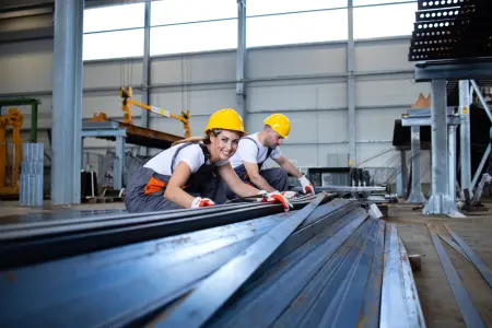 Zwei Arbeiter in Schutzhelmen verarbeiten Metallstücke in einer Fabrikhalle.