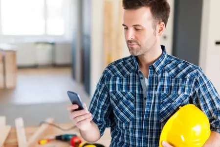 Ein Bauleiter trägt einen Schutzhelm und nutzt ein Smartphone auf einer Baustelle.